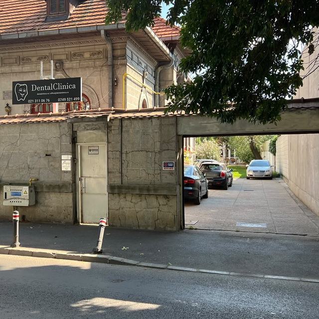 DentalClinica văzută din stradă: firma neagră de deasupra zidului de piatră și, în dreapta, poarta de acces în curte